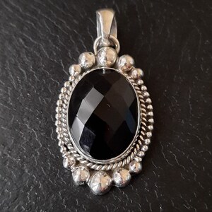 May include: An oval pendant featuring a large, faceted black gemstone set in a silver frame. The border is decorated with a twisted rope pattern and a series of small, polished silver spheres. A silver bail is attached to the top of the pendant.