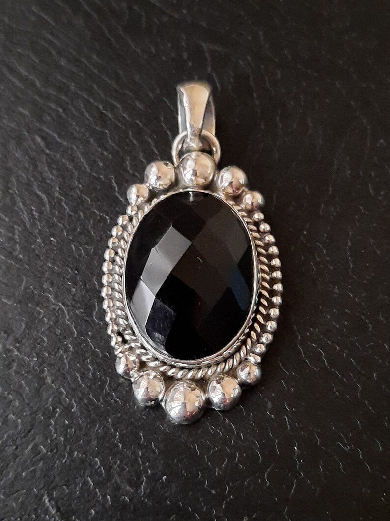 May include: An oval pendant featuring a large, faceted black gemstone set in a silver frame. The border is decorated with a twisted rope pattern and a series of small, polished silver spheres. A silver bail is attached to the top of the pendant.