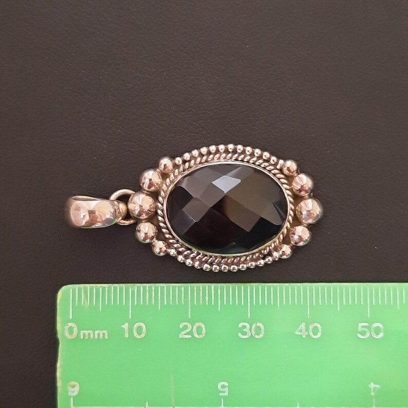 May include: An oval pendant featuring a faceted black gemstone set in a silver frame with a beaded and twisted rope border. The pendant is positioned above a green ruler, showing a width of approximately 1.25 inches from edge to edge.