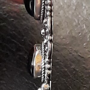 Puede incluir: Un colgante de plata con una piedra preciosa negra y una piedra preciosa transparente. El colgante tiene un dise&ntilde;o detallado con un borde de cuerda y peque&ntilde;as cuentas.