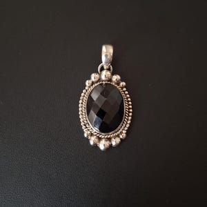 May include: An oval pendant featuring a faceted black gemstone set in a silver frame. The border consists of a twisted rope pattern surrounded by small silver spheres. A polished silver bail is attached to the top for hanging on a chain.