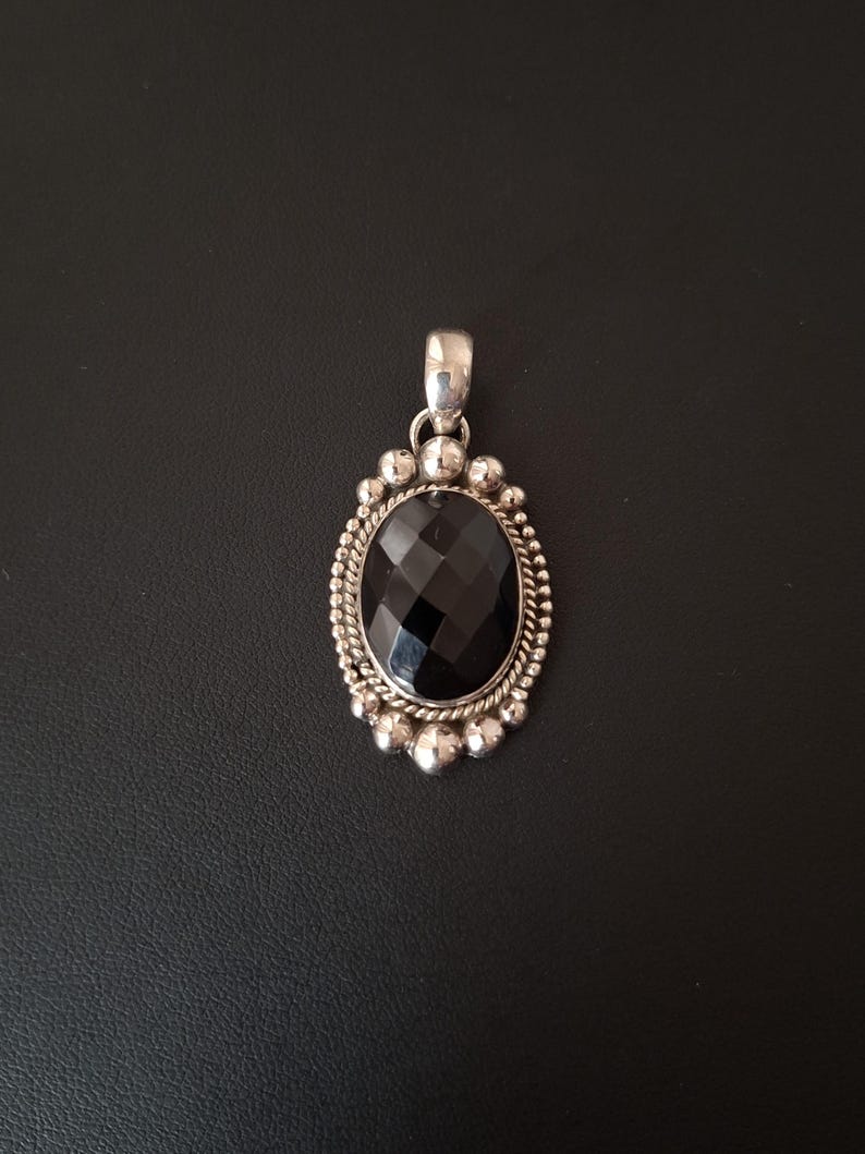 May include: An oval pendant featuring a faceted black gemstone set in a silver frame. The border consists of a twisted rope pattern surrounded by small silver spheres. A polished silver bail is attached to the top for hanging on a chain.