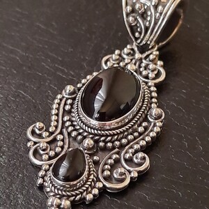 Puede incluir: Un colgante de plata con dos piedras preciosas negras engastadas en un dise&ntilde;o ornamentado. El colgante tiene una anilla en la parte superior para sujetarlo a una cadena.