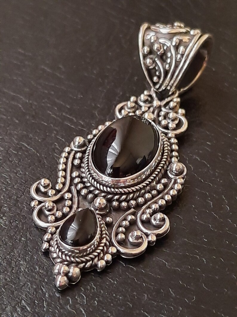 Puede incluir: Un colgante de plata con dos piedras preciosas negras engastadas en un dise&ntilde;o ornamentado. El colgante tiene una anilla en la parte superior para sujetarlo a una cadena.