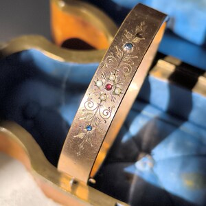 Könnte beinhalten: Ein goldfarbenes, aufklappbares Armreifarmband mit floralen und scrollartigen Gravuren. Das Armband ist mit kleinen, farbigen Edelsteinen, darunter Rot, Blau und Weiß, verziert. Das Armband wird vor dem Hintergrund einer blau samtgefütterten Box präsentiert.