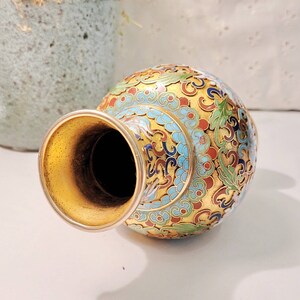 May include: A small, gold-colored vase with a wide mouth and a round body. The vase is decorated with intricate blue, green, red, and white floral patterns.