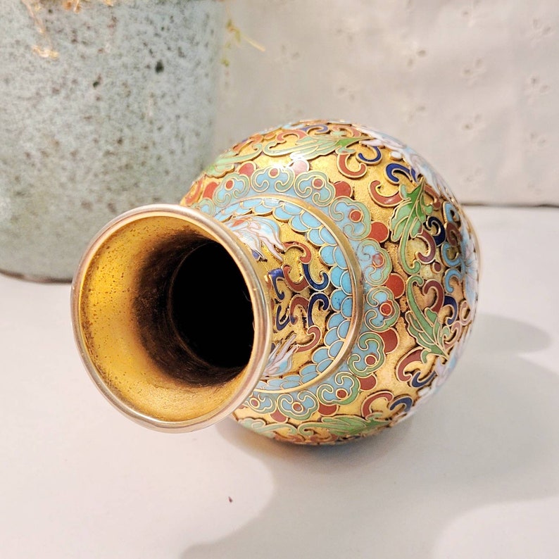 May include: A small, gold-colored vase with a wide mouth and a round body. The vase is decorated with intricate blue, green, red, and white floral patterns.