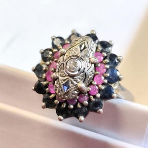 Op de afbeelding: Zilveren ring met een centraal, sierlijk ontwerp met een kleine diamant. De ring is omgeven door een cluster van roze en donkerblauwe edelstenen. De ring is geplaatst tegen een witte achtergrond.