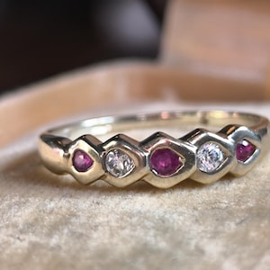 Peut inclure: Une bague en or ornée d'une série de pierres précieuses rouges et claires alternées. Les pierres précieuses sont serties dans un motif répétitif le long de la bande. La bague est présentée sur une surface douce et claire.