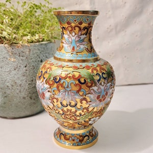 May include: A small, gold-colored vase with a blue, white, and red floral design. The vase is decorated with intricate patterns and has a round body and a flared neck.