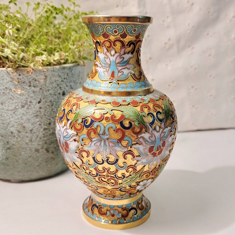 May include: A small, gold-colored vase with a blue, white, and red floral design. The vase is decorated with intricate patterns and has a round body and a flared neck.