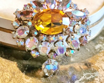 May include: A vintage brooch with a large, oval-shaped amber-colored rhinestone surrounded by smaller, iridescent rhinestones. The brooch has a teardrop-shaped rhinestone dangling from the bottom.