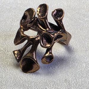 May include: Bronze-toned ring with an organic, abstract design. The ring features multiple rounded, open shapes connected by thin bands, creating a unique, sculptural appearance. The metal has a warm, aged finish, suggesting a vintage or handcrafted style.