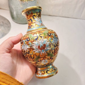May include: A small, gold-colored vase with a blue, white, and red floral pattern. The vase is decorated with intricate details and has a glossy finish.