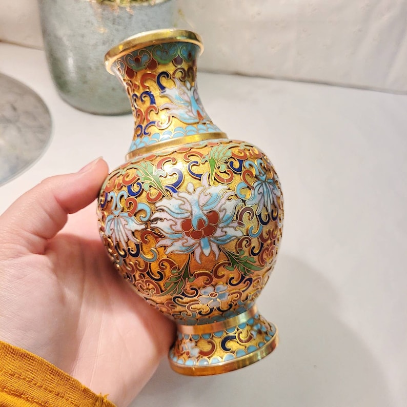 May include: A small, gold-colored vase with a blue, white, and red floral pattern. The vase is decorated with intricate details and has a glossy finish.