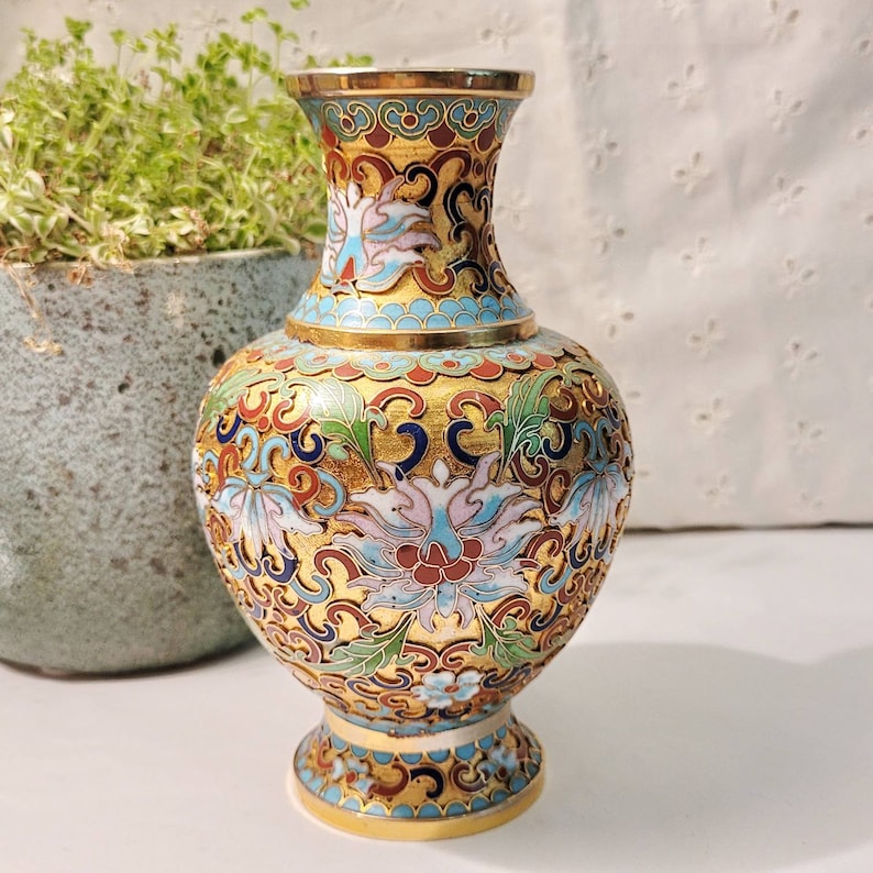 May include: A small, gold-colored vase with a blue and white floral design. The vase has a round body and a flared neck.