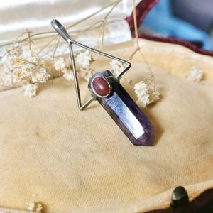 Pode incluir: Um pingente de triângulo prateado com uma ponta de cristal de ametista roxa e um acento de pedra vermelha. O pingente está em uma caixa de joias forrada de veludo.