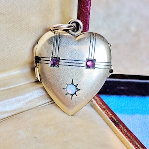 Könnte beinhalten: Ein goldener herzförmiger Anhänger mit einem weißen Opal und zwei roten Edelsteinen. Der Anhänger hat ein zartes schwarzes Design.