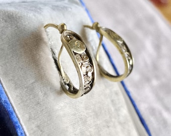 May include: A pair of gold-coloured hoop earrings. One earring has a decorative design with a small circular element. The earrings are displayed inside a blue velvet jewellery box, suggesting a vintage or antique style.