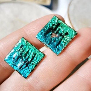 Puede incluir: Un par de pendientes cuadrados con un acabado iridiscente verde y azul. Los pendientes tienen un marco de metal dorado.