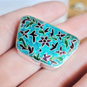 May include: A small, rectangular box with a turquoise enamel finish. The box is decorated with a floral pattern in black, green, and pink.