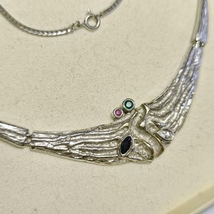 May include: Silver-toned necklace with a textured, organic design. The necklace features a curved centerpiece adorned with a dark blue, a pink, a green, and a clear gemstone. A delicate chain completes the necklace.