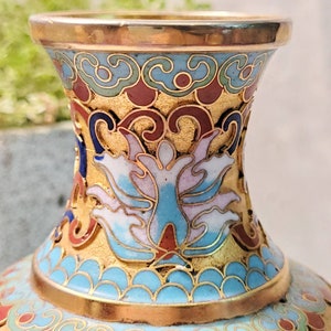 May include: A gold and blue cloisonn&eacute; vase with a floral design. The vase has a wide mouth and a narrow neck. The floral design is in shades of blue, white, and pink.