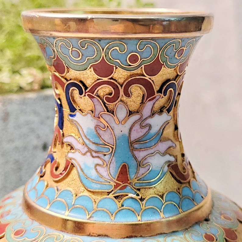 May include: A gold and blue cloisonn&eacute; vase with a floral design. The vase has a wide mouth and a narrow neck. The floral design is in shades of blue, white, and pink.