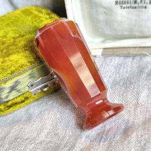 Puede incluir: Un tapón de botella de piedra preciosa rojo-anaranjado facetado. El tapón tiene una base acampanada y un diseño geométrico. El fondo incluye una caja de terciopelo verde y una caja blanca con texto en alemán.