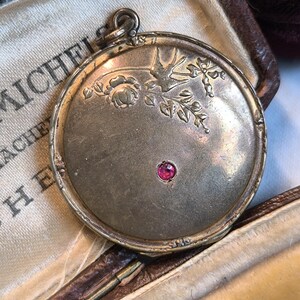 Può includere: Un medaglione d'oro con un designo inciso di un uccello e fiori. Un'unica pietra rossa è incastonata al centro del medaglione.