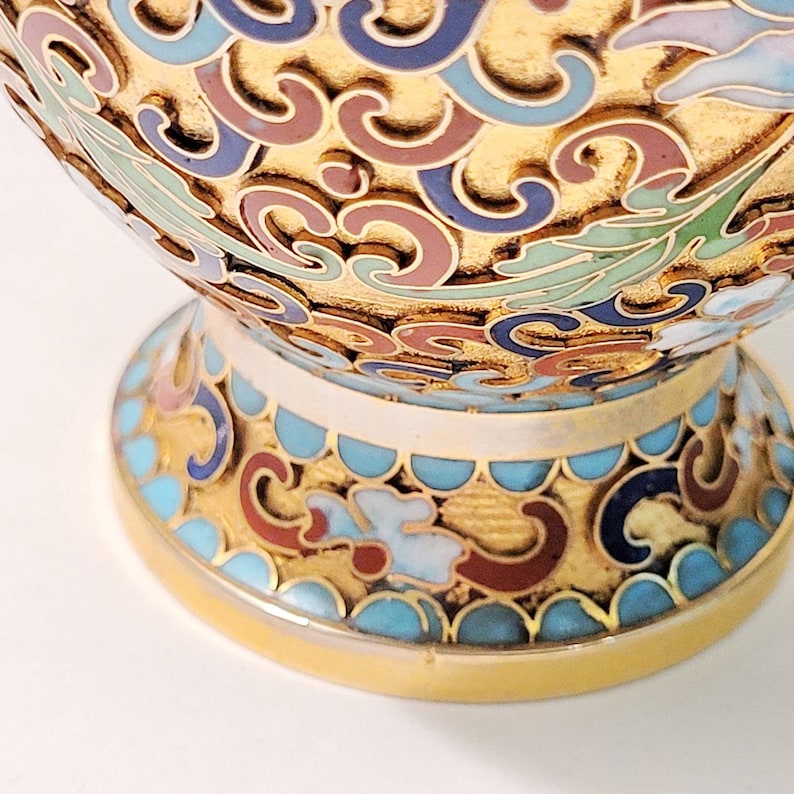 May include: A gold-colored vase with a blue, green, red, and white floral pattern. The vase has a round body and a wide base.