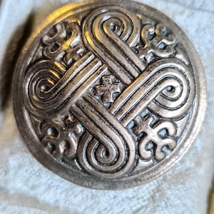 May include: A round, antique-style metal button with an intricate Celtic knot design. The button is a muted bronze color and features raised, swirling patterns and small cross-like details. The button is set against a soft, light-colored background.