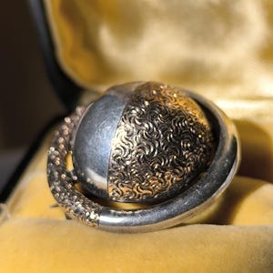 以下が含まれることがあります： シルバーとゴールドのデザインのヴィンテージブローチ。ブローチは、片側にテクスチャ加工されたゴールドパターン、もう片側に滑らかなシルバー仕上げの球状の中心部を備えています。ブローチはシルバーフレームにセットされ、黄色のベルベットの表面に置かれています。