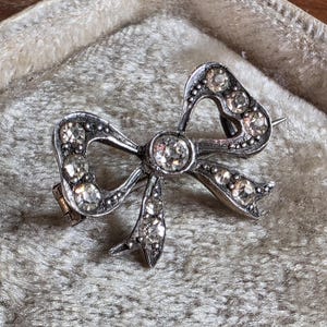 Op de afbeelding: Zilverkleurige strikbroche met heldere, sprankelende edelstenen. De broche heeft een gedetailleerd ontwerp met een centrale steen en kleinere stenen die de strikvorm omlijnen. De speld is geplaatst tegen een zachte, getextureerde achtergrond.