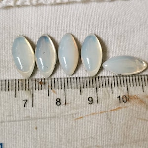 May include: Six iridescent white oval-shaped glass cabochons, measuring approximately 1 cm in length, arranged in a row on a white fabric surface with a ruler for scale.