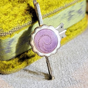 Puede incluir: Una pinza para el pelo de plata con un adorno de flor de tela morada. La flor tiene un borde festoneado y un diseño de espiral.