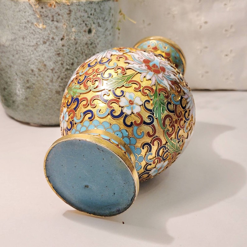 May include: A small, gold-colored vase with a blue, white, and red floral pattern. The vase is decorated with intricate details and has a blue base.