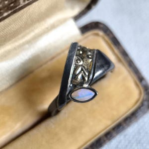 Könnte beinhalten: Ein Silberring mit einem Opalstein. Der Ring hat ein einzigartiges Design mit geometrischen Formen und dekorativen Elementen. Der Opal ist oval und zeigt schillernde Farben. Der Ring ist in einem Vintage-Stil gehalten.