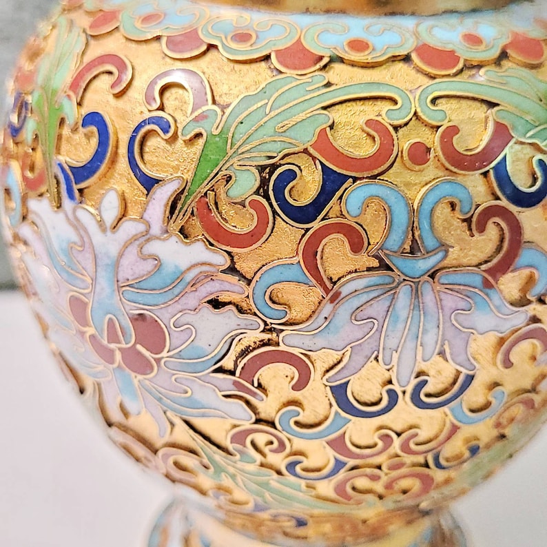 May include: A gold-colored ceramic vase with intricate blue, red, and white floral patterns. The vase has a round body and a narrow neck.