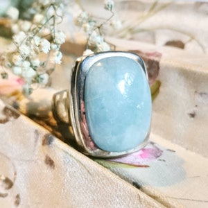 Könnte beinhalten: Ein silberner Ring mit einem großen, hellblauen Aquamarin, der in einer quadratischen Fassung sitzt. Der Ring liegt auf einer weißen Oberfläche mit floralem Muster.