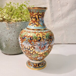 May include: A small, gold-colored vase with a blue, white, and red floral pattern. The vase has a round body and a narrow neck.