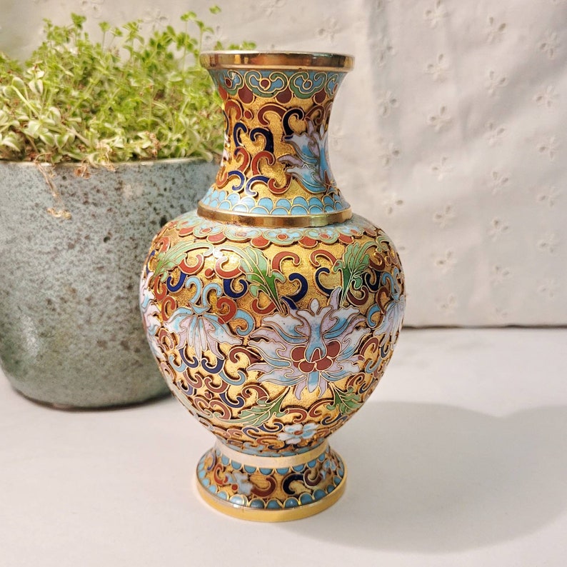 May include: A small, gold-colored vase with a blue, white, and red floral pattern. The vase has a round body and a narrow neck.