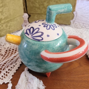 Könnte beinhalten: Eine tealfarbene Teekanne aus Keramik mit einem weißen Deckel, der mit blauen und weißen Blumenmustern verziert ist. Die Teekanne hat einen gelben Ausguss, einen rot-weiß gestreiften Griff und einen tealfarbenen Griff oben.