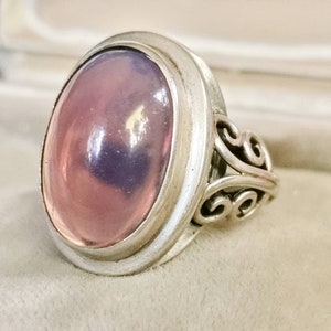 Pode incluir: Um anel de prata com uma grande gema oval rosa engastada em um engaste de bezel. A banda é ornamentada com um design de rolagem.