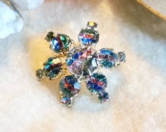 May include: A vintage iridescent flower brooch with a starburst design. The brooch is made of metal and has a gold tone finish. The flower has eight petals and is set with clear, blue, and green iridescent rhinestones.