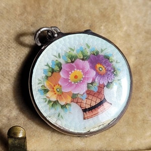 May include: A silver locket with a white enamel background and a hand-painted floral design. The flowers are pink, purple, orange, and yellow with green leaves and blue accents. The locket has a woven basket design at the bottom.