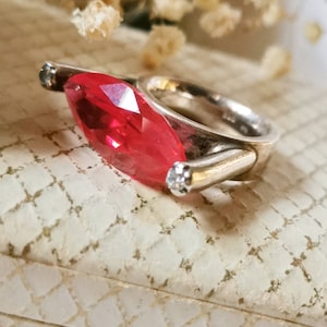 Könnte beinhalten: Ein silberner Ring mit einem großen, roten, markisenförmigen Edelstein und zwei kleineren, klaren Edelsteinen. Der Ring liegt auf einer weißen, strukturierten Oberfläche.