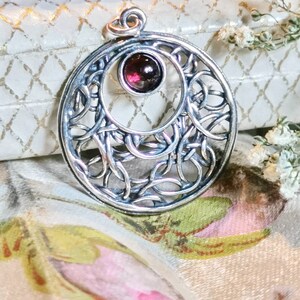 May include: A silver pendant with a delicate, intricate design featuring a circular frame with a central garnet gemstone. The pendant has a Celtic knot-like pattern.