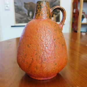 Puede incluir: Un jarrón de cerámica con un esmalte texturizado naranja y verde. El jarrón tiene un cuerpo ancho y redondo y un cuello estrecho con asa.