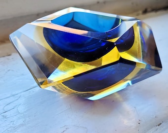 Puede incluir: Un cenicero de vidrio azul y amarillo con un diseño geométrico. El cenicero está hecho de vidrio transparente con un patrón de espiral azul y amarillo.